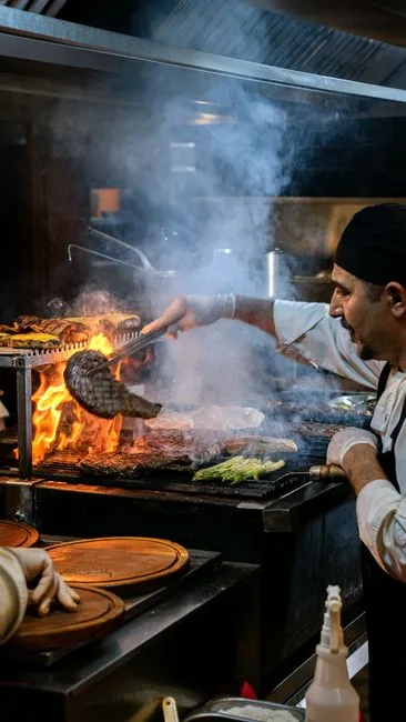 Chef grilling meat with open flames in a commercial kitchen — grease and smoke captured by overhead exhaust hood